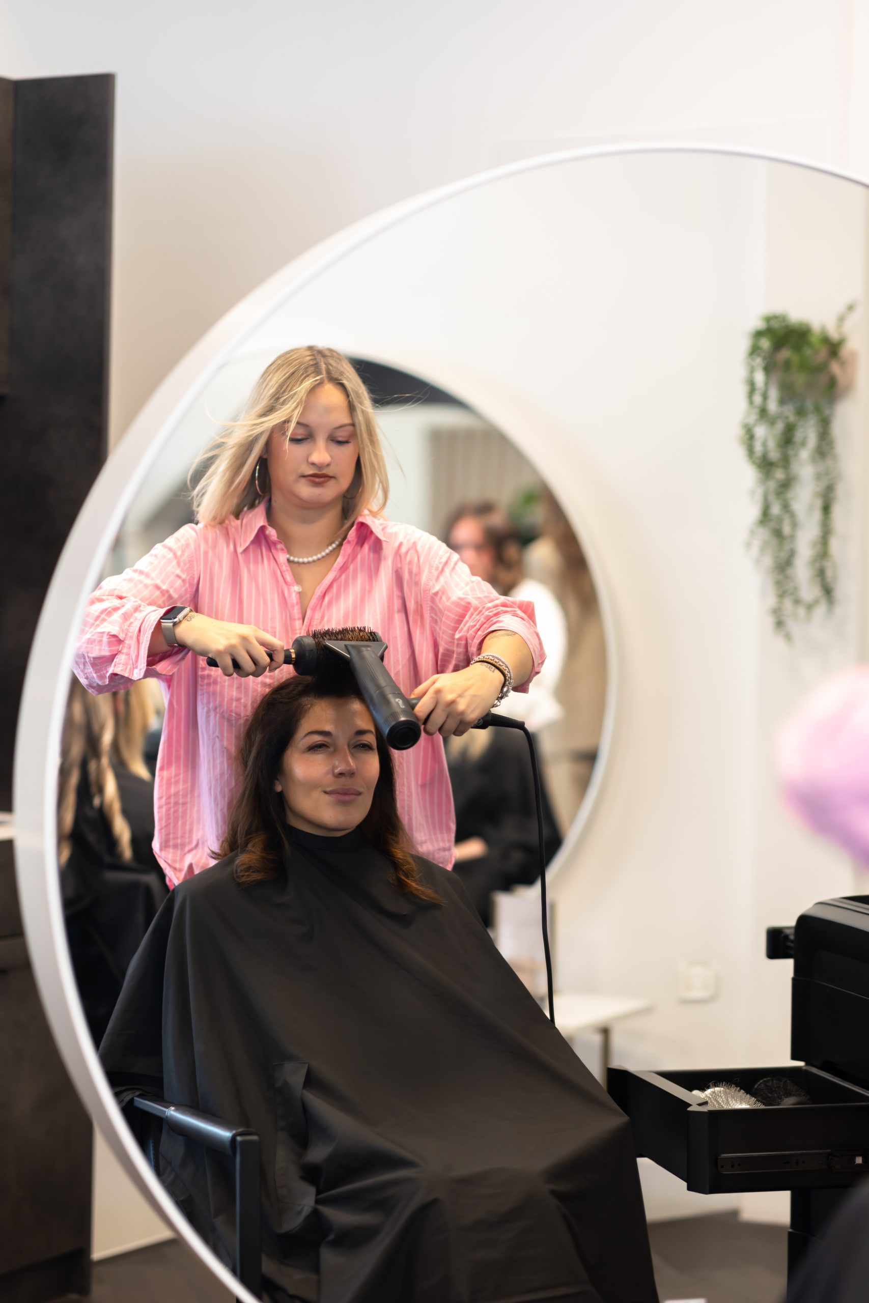 A stylist blow-dries a woman's hair in a salon. The woman, wrapped in a black cape, looks relaxed. A round mirror and plants are visible.
